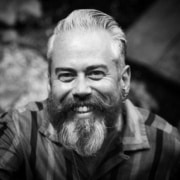 Black-and-white headshot of a person with short light-colored hair, a full beard and mustache, multiple ear piercings, and a patterned button-down shirt, facing the camera.