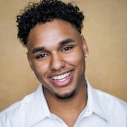 Professional headshot of a person with short curly hair wearing a light-colored collared shirt and stud earrings. The portrait is framed from the shoulders up against a warm beige background with soft studio lighting.