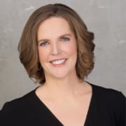 Professional headshot of a person with shoulder-length light brown hair wearing a black V-neck top. The portrait is framed from the shoulders up against a light gray textured background with soft studio lighting.