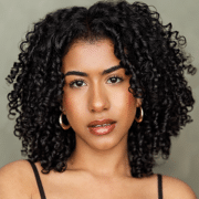 Professional headshot of a person with shoulder-length curly hair wearing hoop earrings and a black sleeveless top. The portrait is framed from the shoulders up against a soft neutral background with studio lighting.