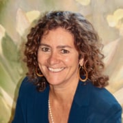 Professional headshot of a person with shoulder-length curly hair wearing a blue blazer, hoop earrings, and a pearl necklace. The portrait is framed from the chest up against a softly painted floral background.