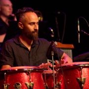 Musician performs on red conga drums during a live concert. Microphones surround the percussion setup, while additional musicians and stage equipment are visible in the softly lit background under colorful stage lighting.