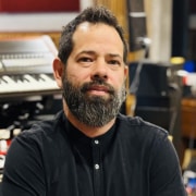 Professional portrait of a person with a beard wearing a black buttoned shirt. The image is photographed in a recording studio environment, with keyboards, audio equipment, and instruments visible in the softly blurred background.
