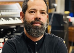 Professional portrait of a person with a beard wearing a black buttoned shirt. The image is photographed in a recording studio environment, with keyboards, audio equipment, and instruments visible in the softly blurred background.