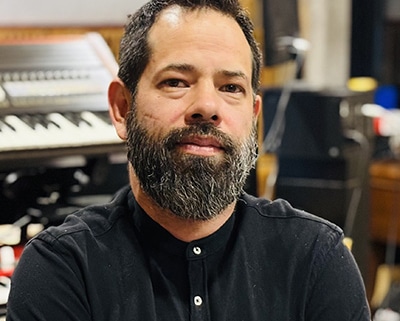 Professional portrait of a person with a beard wearing a black buttoned shirt. The image is photographed in a recording studio environment, with keyboards, audio equipment, and instruments visible in the softly blurred background.