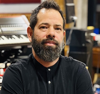 Professional portrait of a person with a beard wearing a black buttoned shirt. The image is photographed in a recording studio environment, with keyboards, audio equipment, and instruments visible in the softly blurred background.