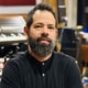 Professional portrait of a person with a beard wearing a black buttoned shirt. The image is photographed in a recording studio environment, with keyboards, audio equipment, and instruments visible in the softly blurred background.