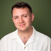 Professional headshot of a person with short dark hair wearing a white collared shirt. The portrait is framed from the shoulders up against a soft green background with even studio lighting.