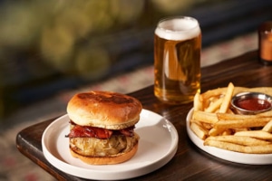 Gourmet burger topped with caramelized onions and bacon, served with French fries, ketchup, and a pint of beer.