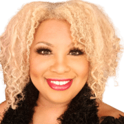 Professional headshot of a person with light-colored curly hair wearing a dark textured top. The portrait is tightly cropped against a white background with bright, even lighting.