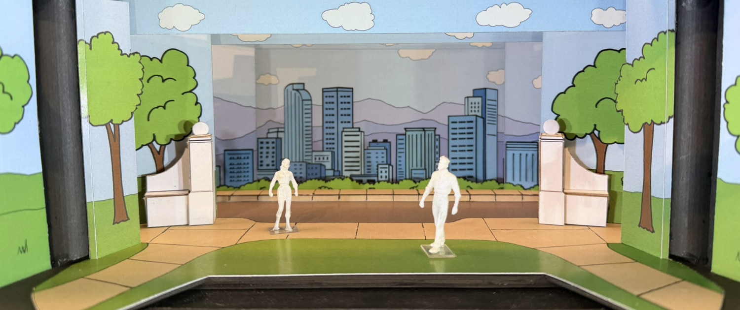 Scale model rendering of a stage set for Don’t Let the Pigeon Drive the Bus featuring a colorful cartoon-style city park. Painted flats depict green trees, blue sky, clouds, and a city skyline with mountains in the background. White architectural elements frame the center stage area, while two miniature figure models stand on a curved green-and-tan playing space in the foreground. The model is displayed inside a dark wooden presentation box.