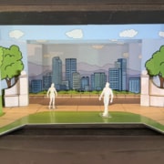 Scale model rendering of a stage set for Don’t Let the Pigeon Drive the Bus featuring a colorful cartoon-style city park. Painted flats depict green trees, blue sky, clouds, and a city skyline with mountains in the background. White architectural elements frame the center stage area, while two miniature figure models stand on a curved green-and-tan playing space in the foreground. The model is displayed inside a dark wooden presentation box.
