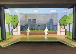 Scale model rendering of a stage set for Don’t Let the Pigeon Drive the Bus featuring a colorful cartoon-style city park. Painted flats depict green trees, blue sky, clouds, and a city skyline with mountains in the background. White architectural elements frame the center stage area, while two miniature figure models stand on a curved green-and-tan playing space in the foreground. The model is displayed inside a dark wooden presentation box.