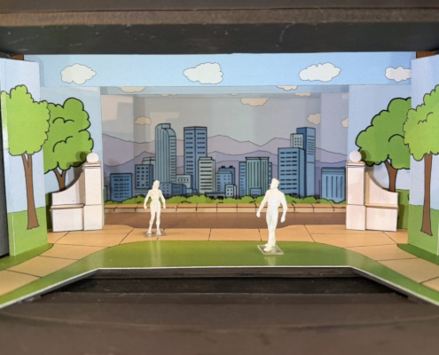 Scale model rendering of a stage set for Don’t Let the Pigeon Drive the Bus featuring a colorful cartoon-style city park. Painted flats depict green trees, blue sky, clouds, and a city skyline with mountains in the background. White architectural elements frame the center stage area, while two miniature figure models stand on a curved green-and-tan playing space in the foreground. The model is displayed inside a dark wooden presentation box.