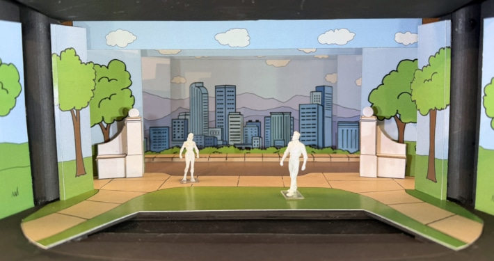 Scale model rendering of a stage set for Don’t Let the Pigeon Drive the Bus featuring a colorful cartoon-style city park. Painted flats depict green trees, blue sky, clouds, and a city skyline with mountains in the background. White architectural elements frame the center stage area, while two miniature figure models stand on a curved green-and-tan playing space in the foreground. The model is displayed inside a dark wooden presentation box.