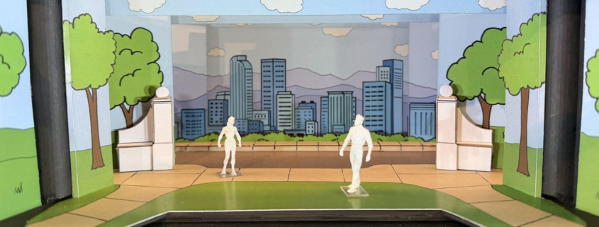 Scale model rendering of a stage set for Don’t Let the Pigeon Drive the Bus featuring a colorful cartoon-style city park. Painted flats depict green trees, blue sky, clouds, and a city skyline with mountains in the background. White architectural elements frame the center stage area, while two miniature figure models stand on a curved green-and-tan playing space in the foreground. The model is displayed inside a dark wooden presentation box.
