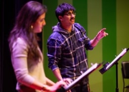 Participant speaks from a script at a music stand during a staged reading, while another participant stands nearby on a theater stage illuminated with green and purple lighting.