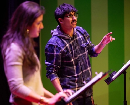 Participant speaks from a script at a music stand during a staged reading, while another participant stands nearby on a theater stage illuminated with green and purple lighting.
