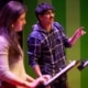 Participant speaks from a script at a music stand during a staged reading, while another participant stands nearby on a theater stage illuminated with green and purple lighting.