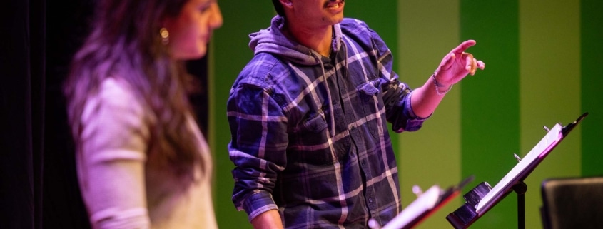 Participant speaks from a script at a music stand during a staged reading, while another participant stands nearby on a theater stage illuminated with green and purple lighting.