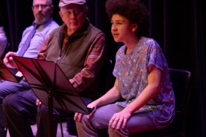 Participants sit onstage during a playwriting workshop reading, with a young reader speaking from a seated position beside script stands and fellow readers.