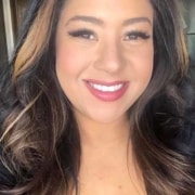 Professional headshot of a person with long dark hair wearing a dark top. The portrait is closely cropped against a softly blurred indoor background, with natural lighting and the subject facing the camera.