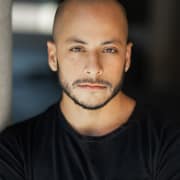 Professional headshot of a person with a shaved head and a neatly trimmed beard, wearing a black shirt. The portrait is framed from the chest up against a softly blurred dark background with natural-looking light.
