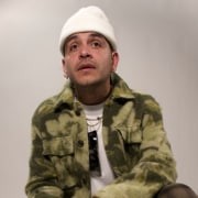 Professional portrait of a person wearing a white knit beanie, layered necklaces, and a green plaid overshirt over a white T-shirt. The image is framed from the waist up against a light studio background with soft lighting.