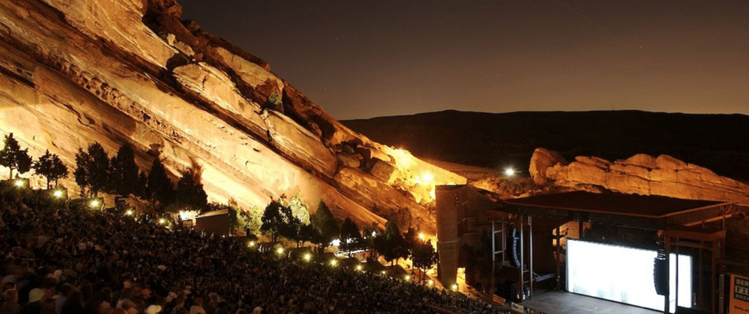 Nighttime view of Red Rocks Amphitheatre, with a large audience seated among towering illuminated red sandstone formations surrounding an outdoor stage.