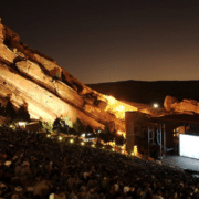Nighttime view of Red Rocks Amphitheatre, with a large audience seated among towering illuminated red sandstone formations surrounding an outdoor stage.