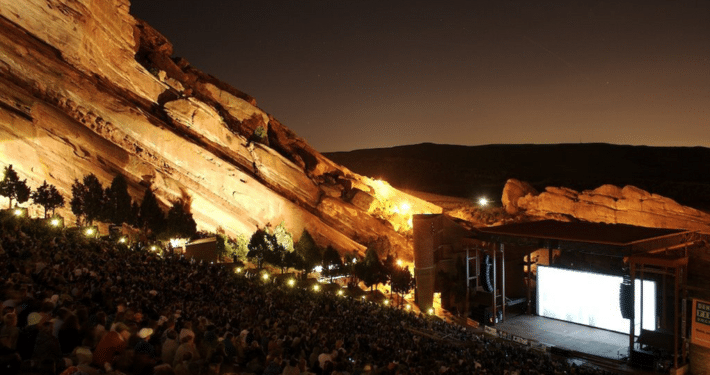 Nighttime view of Red Rocks Amphitheatre, with a large audience seated among towering illuminated red sandstone formations surrounding an outdoor stage.