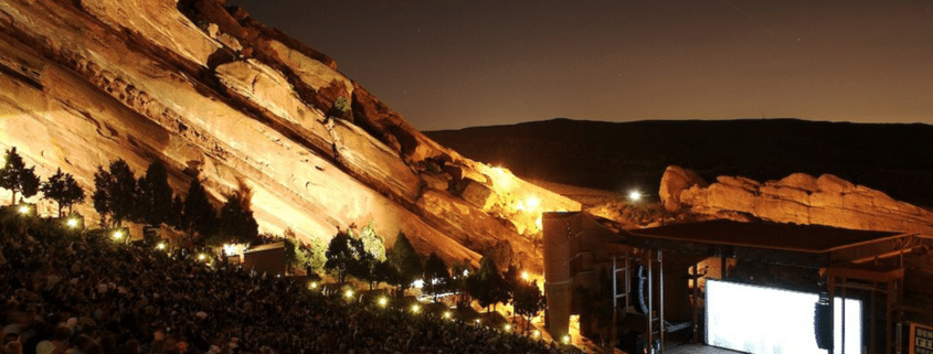 Nighttime view of Red Rocks Amphitheatre, with a large audience seated among towering illuminated red sandstone formations surrounding an outdoor stage.