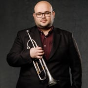 Professional portrait of a person wearing glasses, a dark suit jacket, and a burgundy shirt while holding a silver trumpet. The image is framed from the waist up against a dark textured studio background with soft lighting.