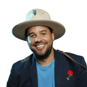 Professional portrait of a person wearing a wide-brimmed light-colored hat, blue shirt, and dark blazer with a rose-shaped lapel pin. The image is framed from the chest up against a light background.