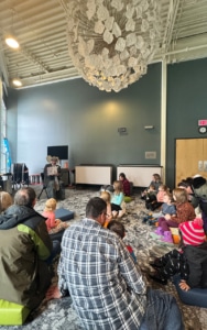 Families and children gather for an interactive storytime program, listening to costumed performers read books in a children's library space.