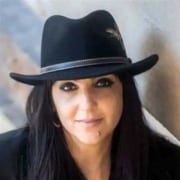 rofessional portrait of a person with long dark hair wearing a black hat and dark clothing. The image is closely cropped against a softly blurred neutral background.