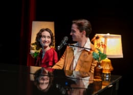 Two performers seated together at a grand piano share a glance beside a microphone, surrounded by soft lamp light and floral accents during a live stage performance.