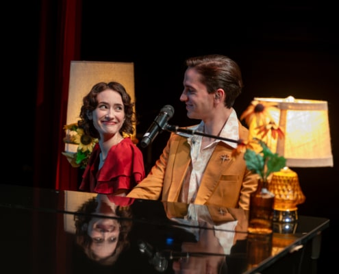 Two performers seated together at a grand piano share a glance beside a microphone, surrounded by soft lamp light and floral accents during a live stage performance.