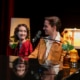 Two performers seated together at a grand piano share a glance beside a microphone, surrounded by soft lamp light and floral accents during a live stage performance.