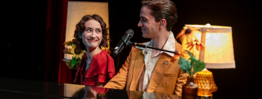 Two performers seated together at a grand piano share a glance beside a microphone, surrounded by soft lamp light and floral accents during a live stage performance.