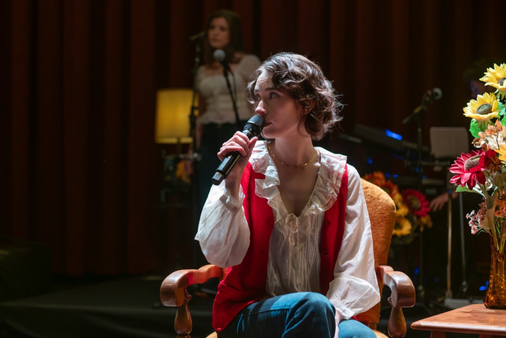Performer seated in an armchair sings into a handheld microphone onstage, framed by sunflowers, warm lighting, and musicians visible in the background.