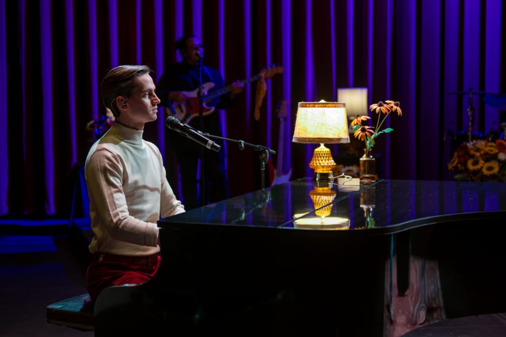 Performer seated at a glossy black grand piano sings into a microphone on a warmly lit stage, with a guitarist in the background and decorative lamps and flowers creating an intimate concert setting.