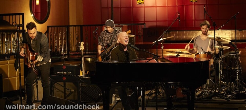 Band performing in a recording studio, with a vocalist seated at a grand piano while other musicians play guitar, bass, and drums under warm ambient lighting.
