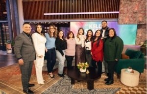 A group of ten people standing together on a television studio set in front of a colorful backdrop and seating area.