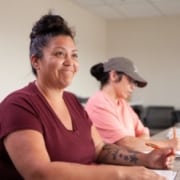 A participant seated at a classroom table during a training session, with another participant taking notes in the background.