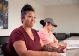 A participant seated at a classroom table during a training session, with another participant taking notes in the background.