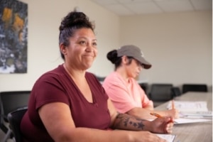 A participant seated at a classroom table during a training session, with another participant taking notes in the background.