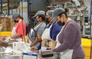 Participants work at a packaging station in an industrial workspace, handling boxed products and equipment.