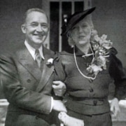 Black-and-white photograph of two formally dressed people standing arm in arm outdoors. One wears a dark suit with a boutonniere, while the other wears a dark dress, hat, floral corsage, and long necklace.