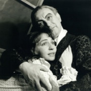 Black-and-white theatrical production photograph of two performers in period costumes embracing onstage, with one performer standing behind and wrapping both arms around the other beneath dramatic stage lighting.